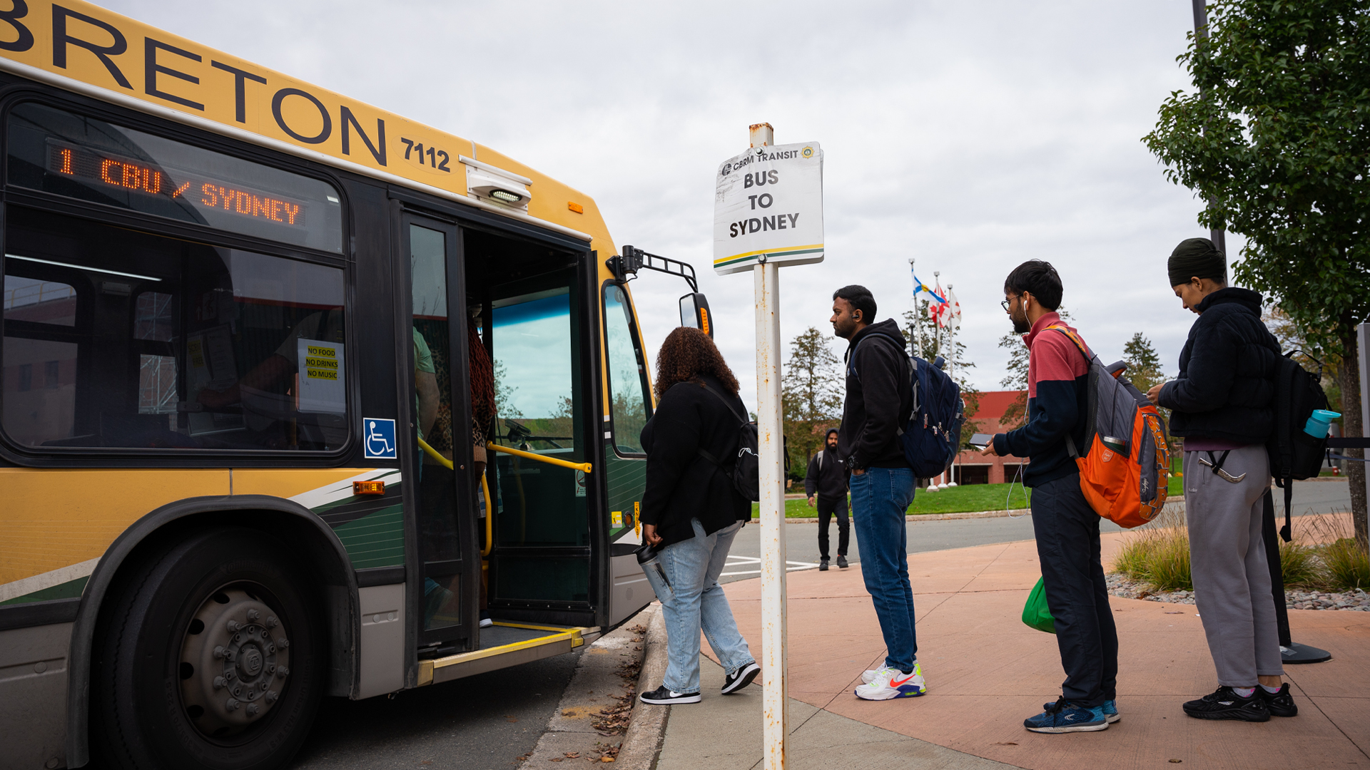Four young people, most of them South Asians, stand in a line at a bus stop with a small square white sign that says “Bus to Sydney”. The woman at the front of the line is about to step on a bus.