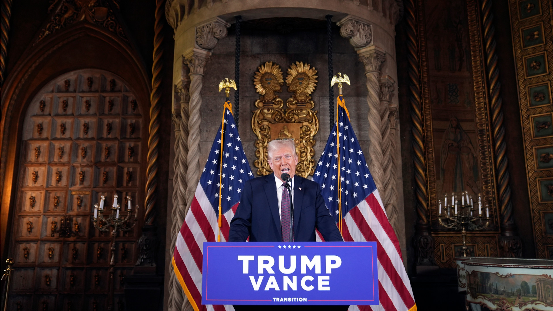 Donald Trump speaks in front of a “Trump-Vance” podium with the dark, garish neogothic décor of Mar-a-Lago behind him.