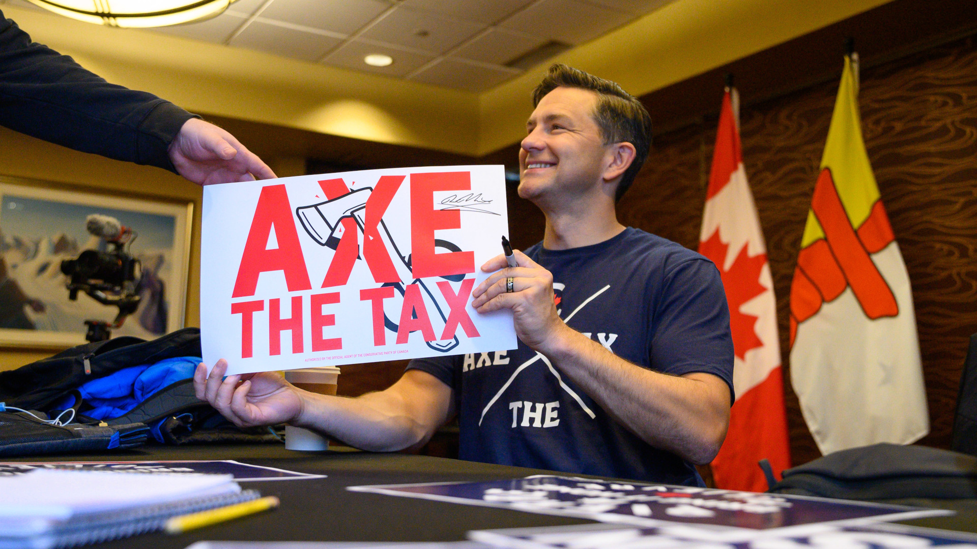 Poilievre smiles as he holds a white and red sign that says “axe the tax,” with a black sharpie in one hand. He’s seated at a table with a couple of flags draped behind him. He’s in a black t-shirt with the “axe the tax” slogan on it.
