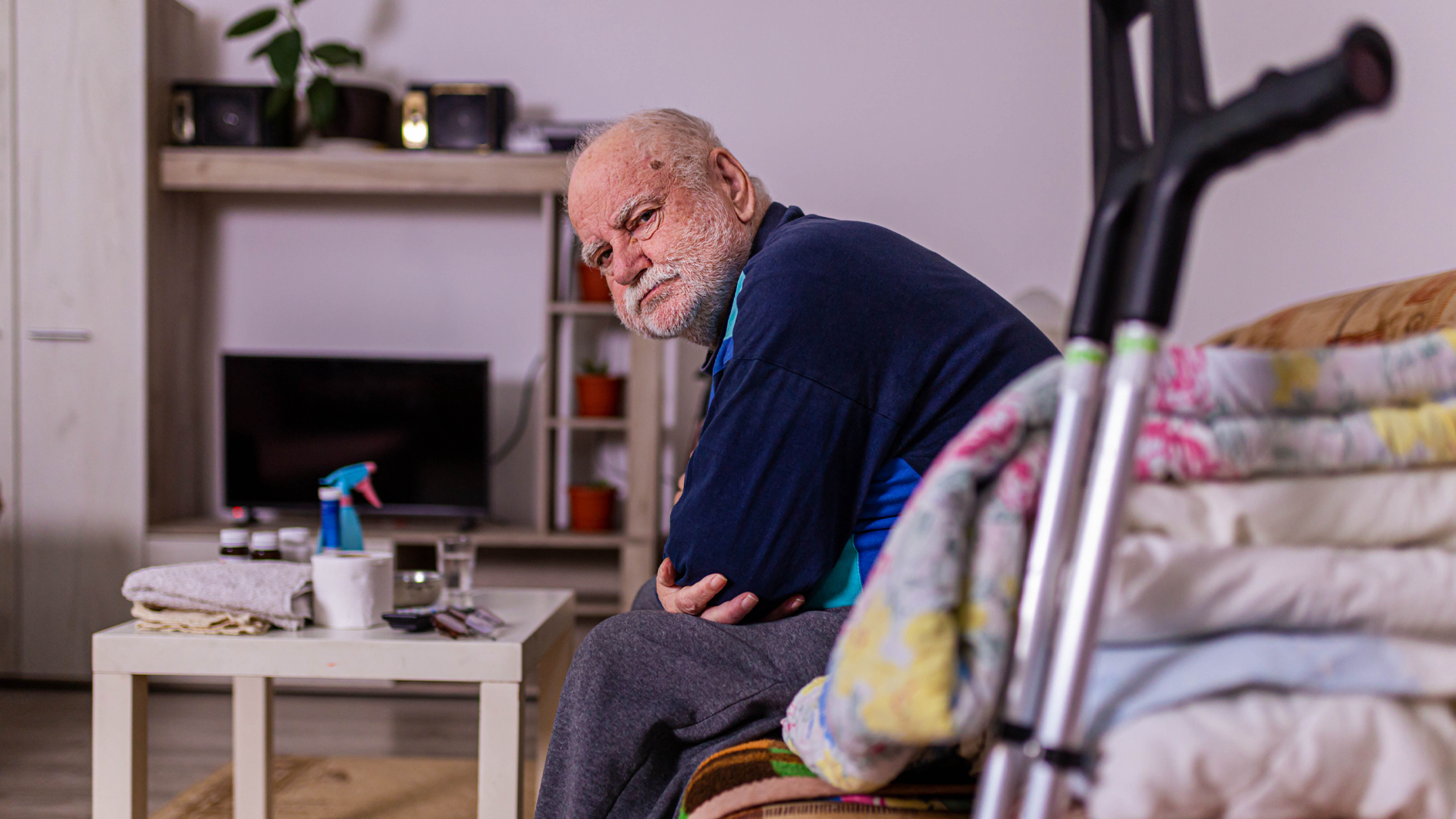 Older white man sits on a sofa as he leans forward with folded arms. A set of crutches lean against the furniture. The room is Spartan with a small white square plastic table covered in items like a roll of toilet paper and a small spray bottle. A stack of folded linens sit at the end of the bed.