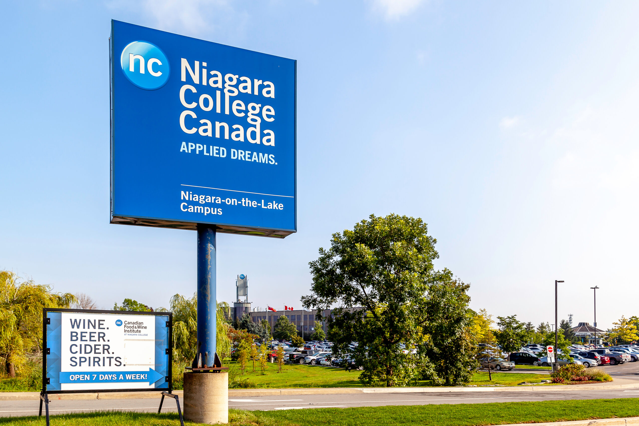 A tall blue sign says “Niagara College Canada. Applied dreams.” A smaller temporary white sign says Wine, beer, cider, spirits. Open 7 days a week!” In the distance is a parking lot and a long, low-rise building. Lots of trees around.