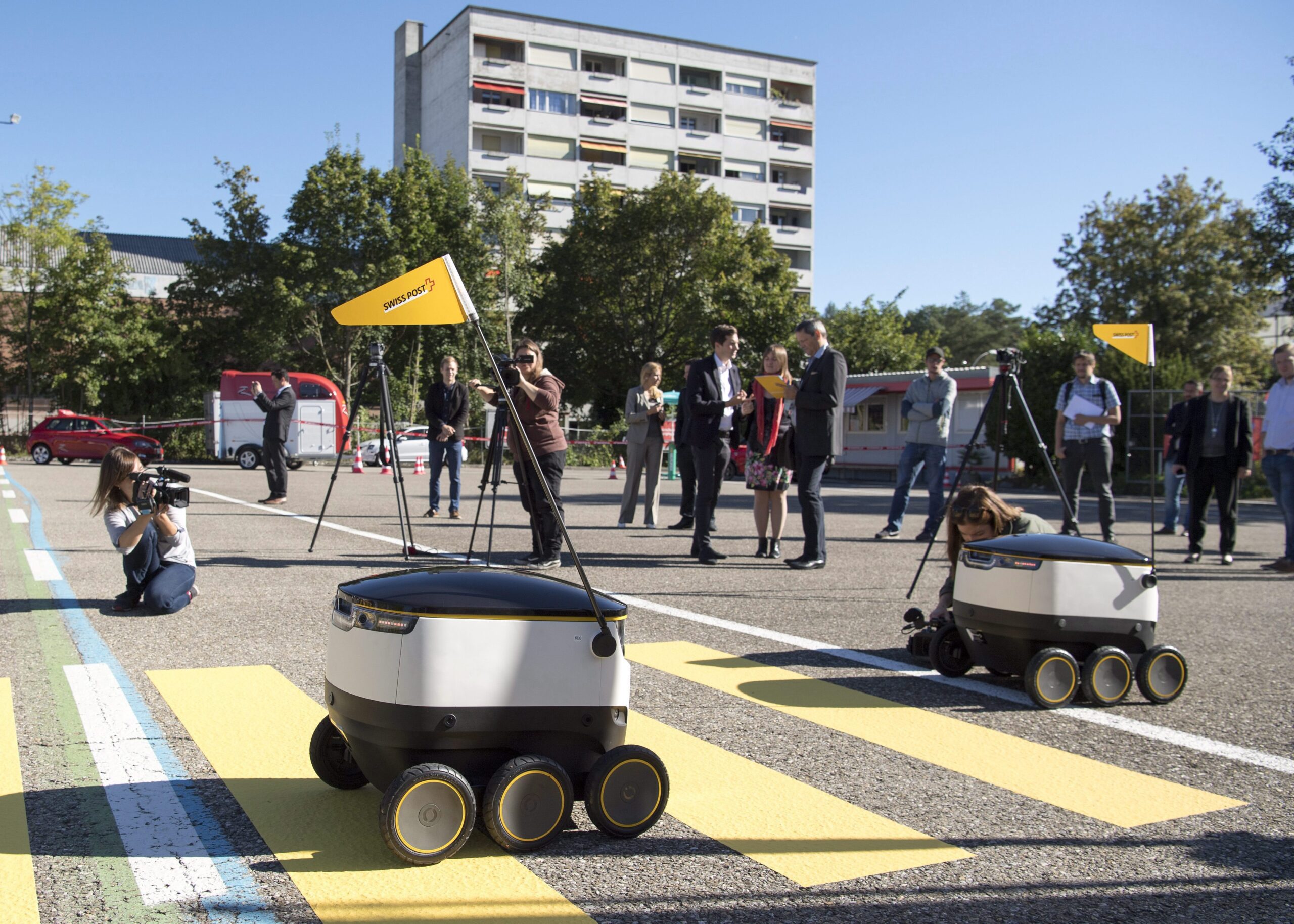 Two of the robots are crossing a crosswalk while several photographers shoot them as a handful of men and women stand around talking. The robots are small, compact containers with curved sides and three wheels on two sides. Each unit has a yellow triangular flag on a thin metal pole that extends a few feet above the robot bodies. The street is lined with trees. A beige concrete apartment building with balconies is in the distance.