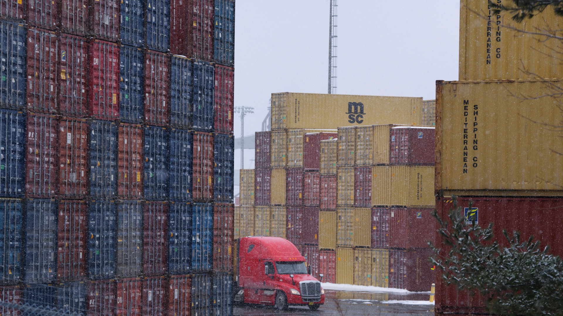 Massive stacks of red, blue and yellow containers are seen through driving snow with a red tractor trailer driving among them.