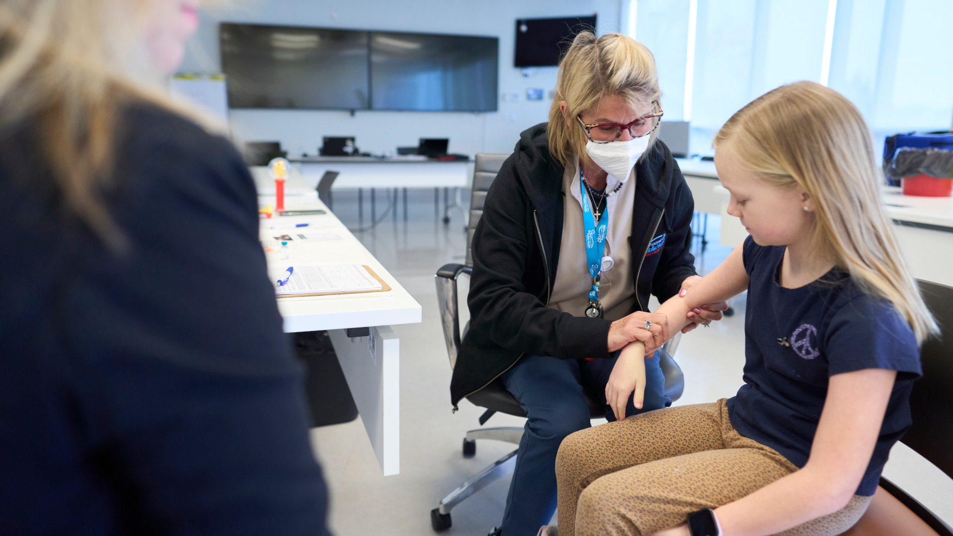 A nurse is holding the arm of a young blond girl in a clinic while another adult looks on. 