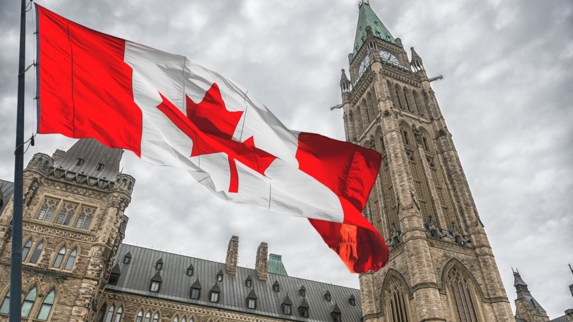 Un drapeau du Canada flotte au-dessus du Parlement. Des nuages gris assombrissent le ciel.