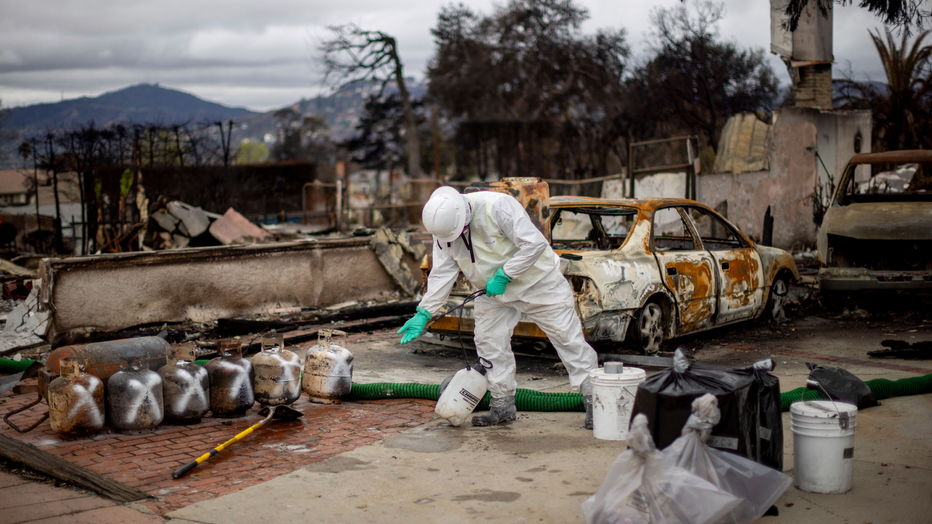 A worker clad in a white jumpsuit and hardhat sprays their gloved hand. The burnt-out remains of several vehicles and a neighbourhood are in the background. A line of barbeque propane tanks are maked with white Xs.