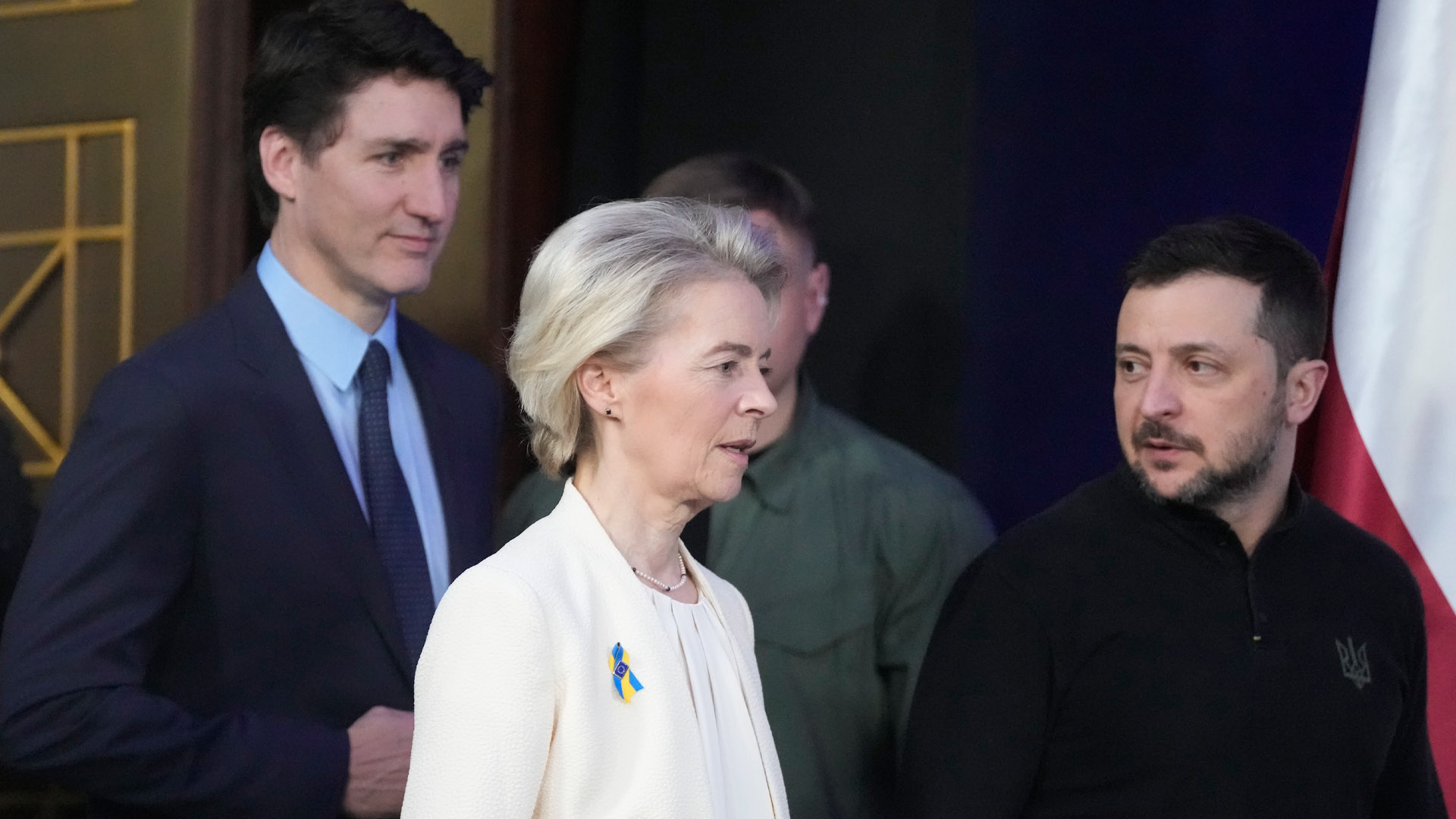 Ukrainian President Volodymyr Zelenskyy in a black sweater looks back at European Commission President Ursula von der Leyen in a cream coloured jacket and blouse while Justin Trudeau follows behind in a blue business suit.