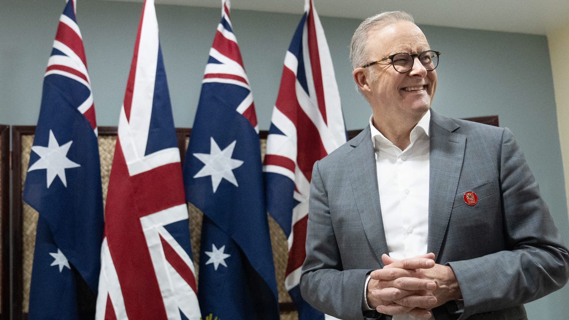 Albanese is standing next to a row of four draped flags, with two Aussie flags alternating with the Union Jack. He is smiling broadly as he looks to the right. His hands are folded in front of his chest. He wears a grey jacket and a white shirt with an open collar.