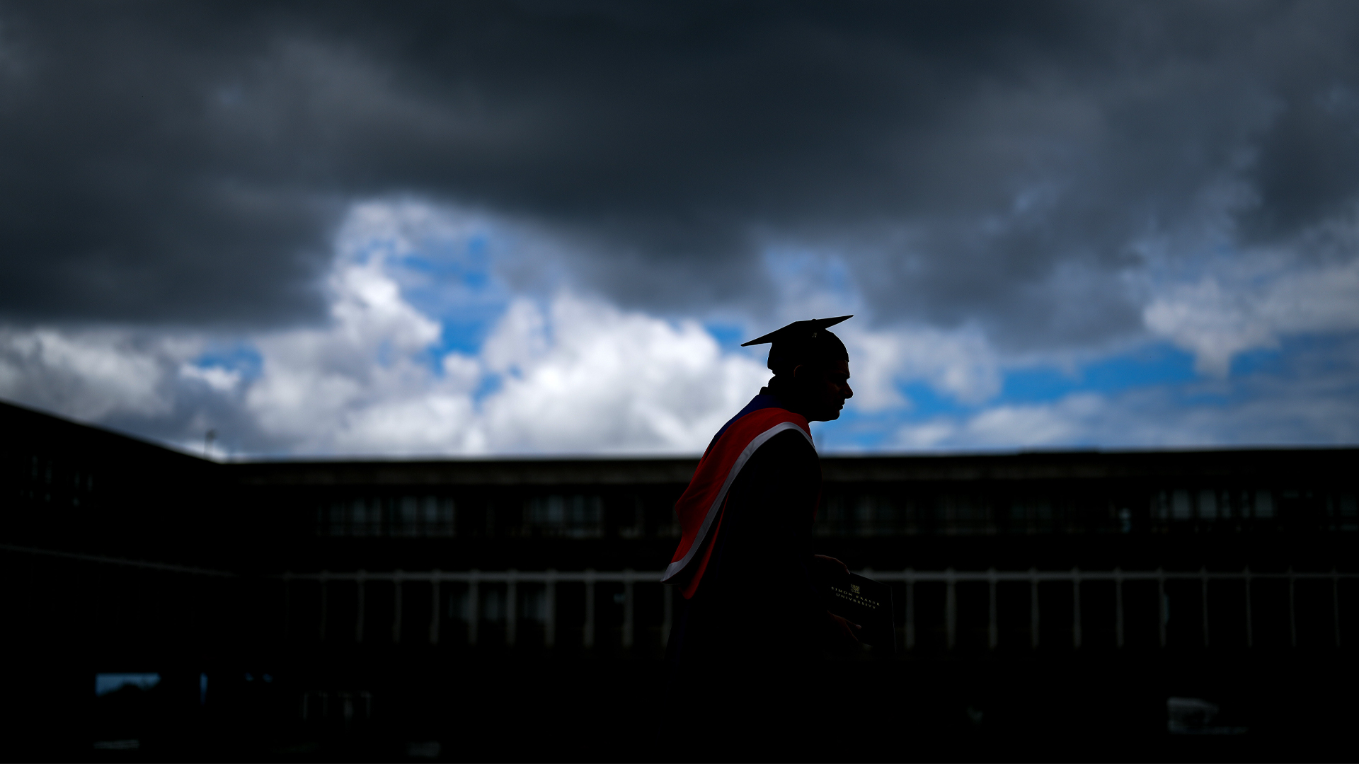 The student wearing a gown and cap is seen in silhouette against a cloudy sky with patches of blue.