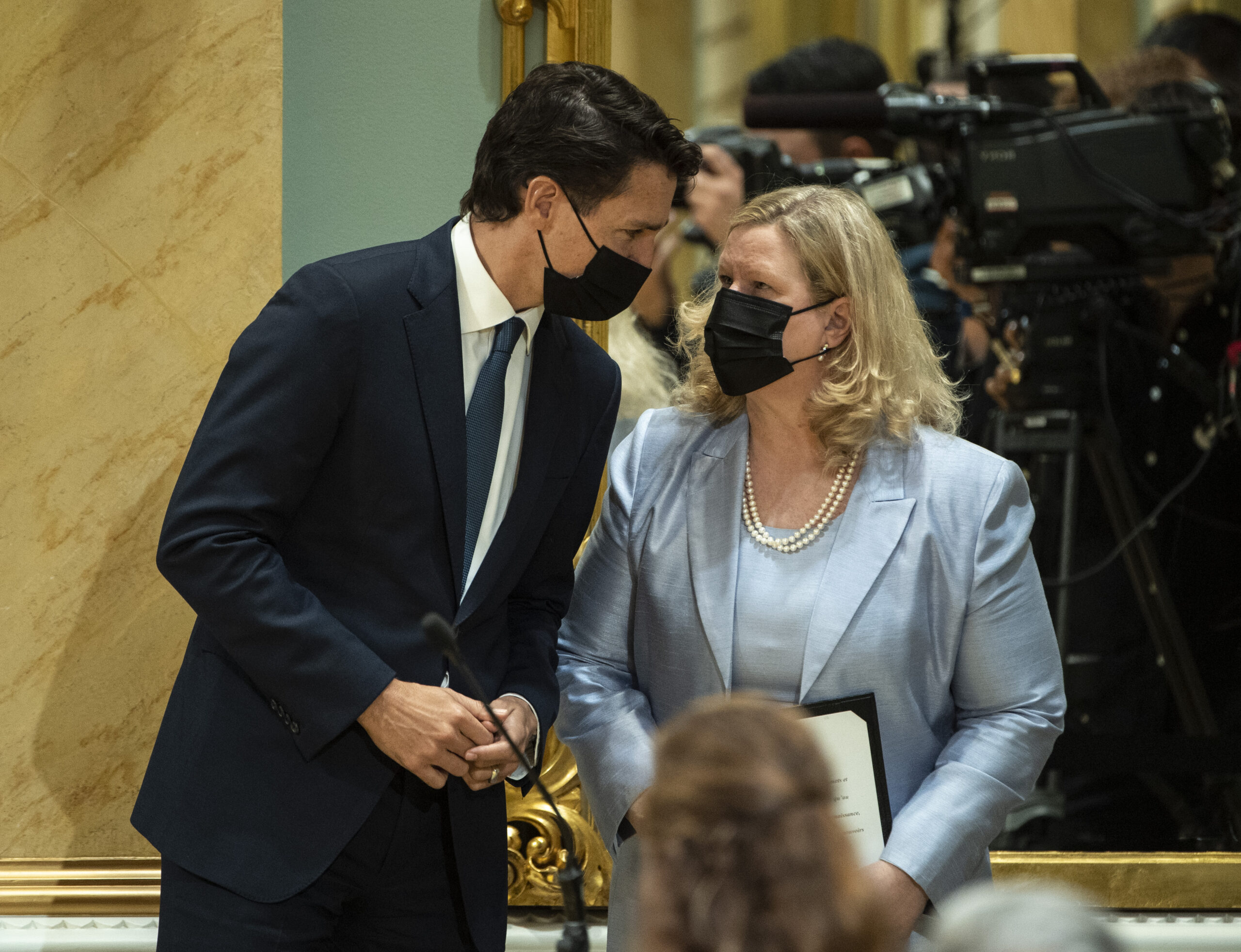 Trudeau is leaning in to speak with Charette. In the background is a bank of television cameras and cameramen. He’s in a black suit; she’s in a robin’s-egg blue suit with pearls. Both are wearing black medical masks.
