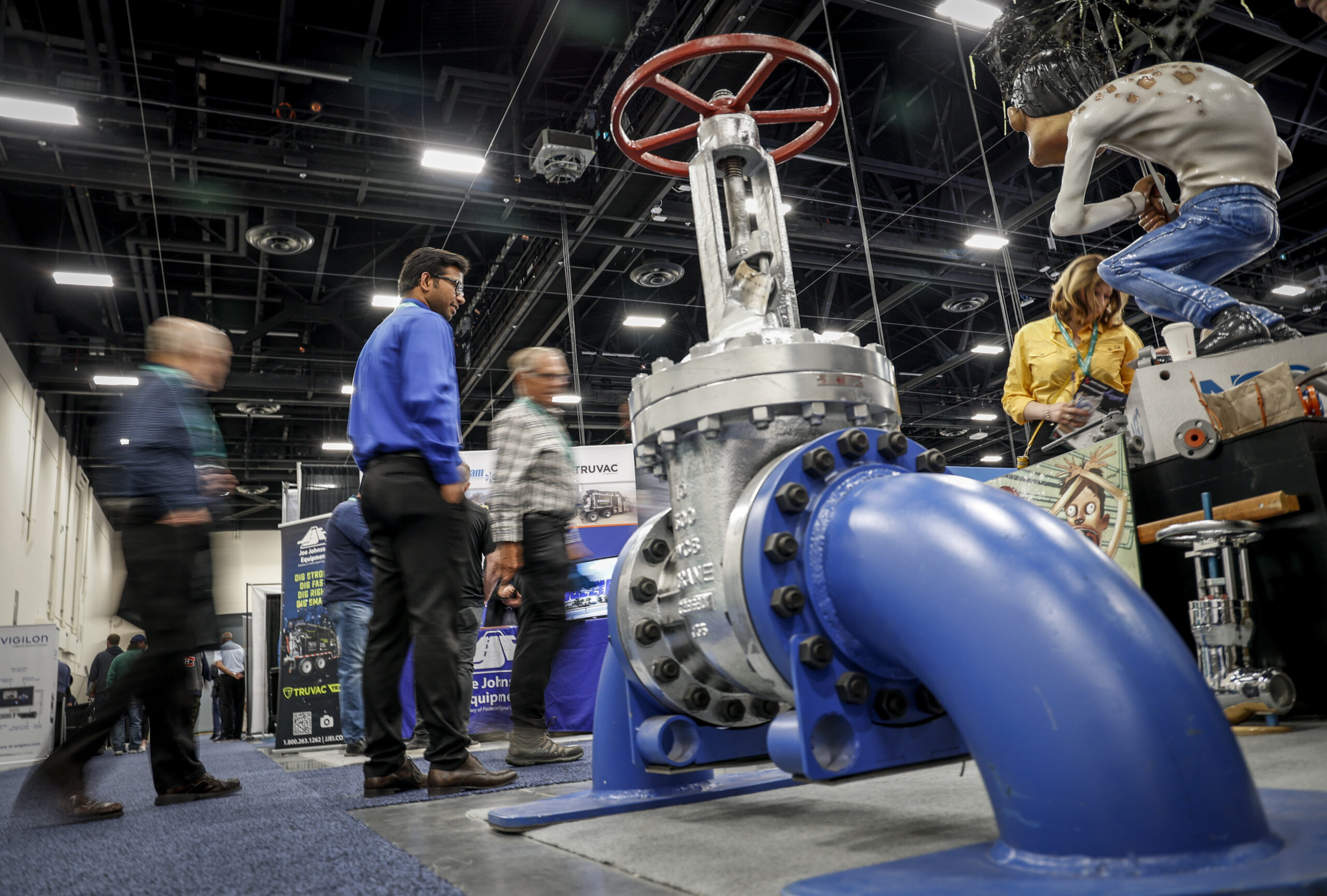 Men and women mill around large pieces of equipment including one made of pipes connected to a valve.
