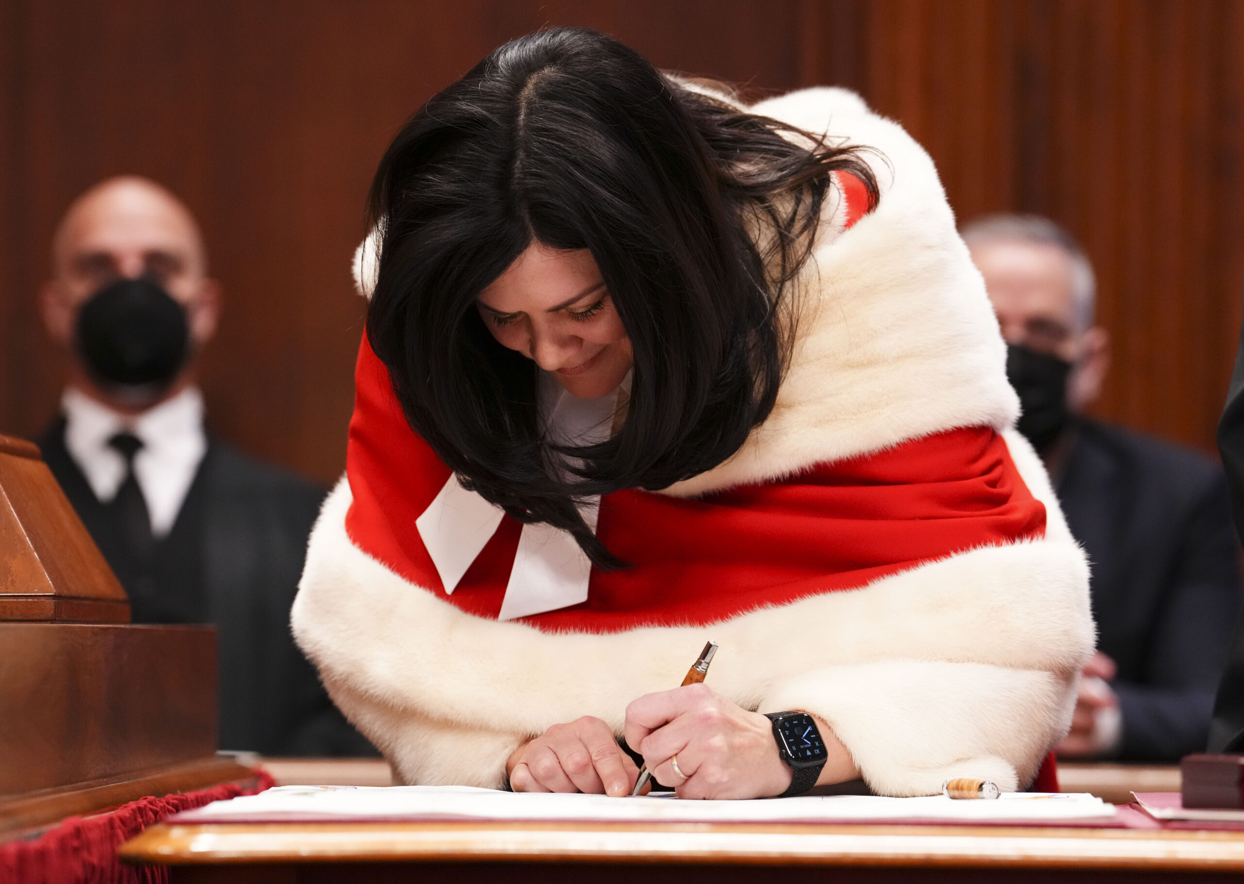A smiling O’Bonsawin is leaning over a surface as she writes on paper. She is dressed in the red robes of her profession, red with white fur trim. Behind her sit two men in black suits and black COVID masks. O’Bonsawin’s black hair falls freely, framing her face and chin.
