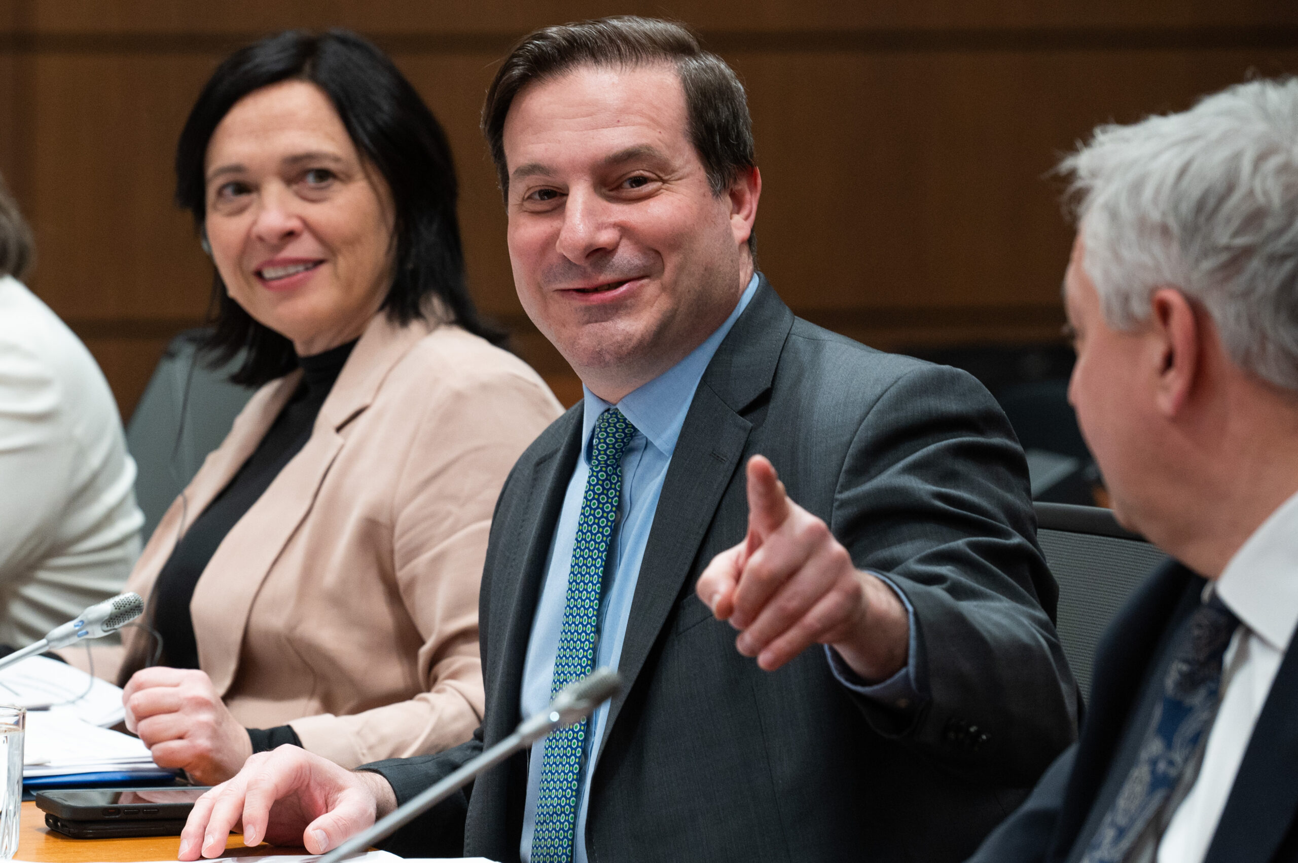 Mendicino is smiling as he points a finger at the camera. He is seated at a long table with other people.