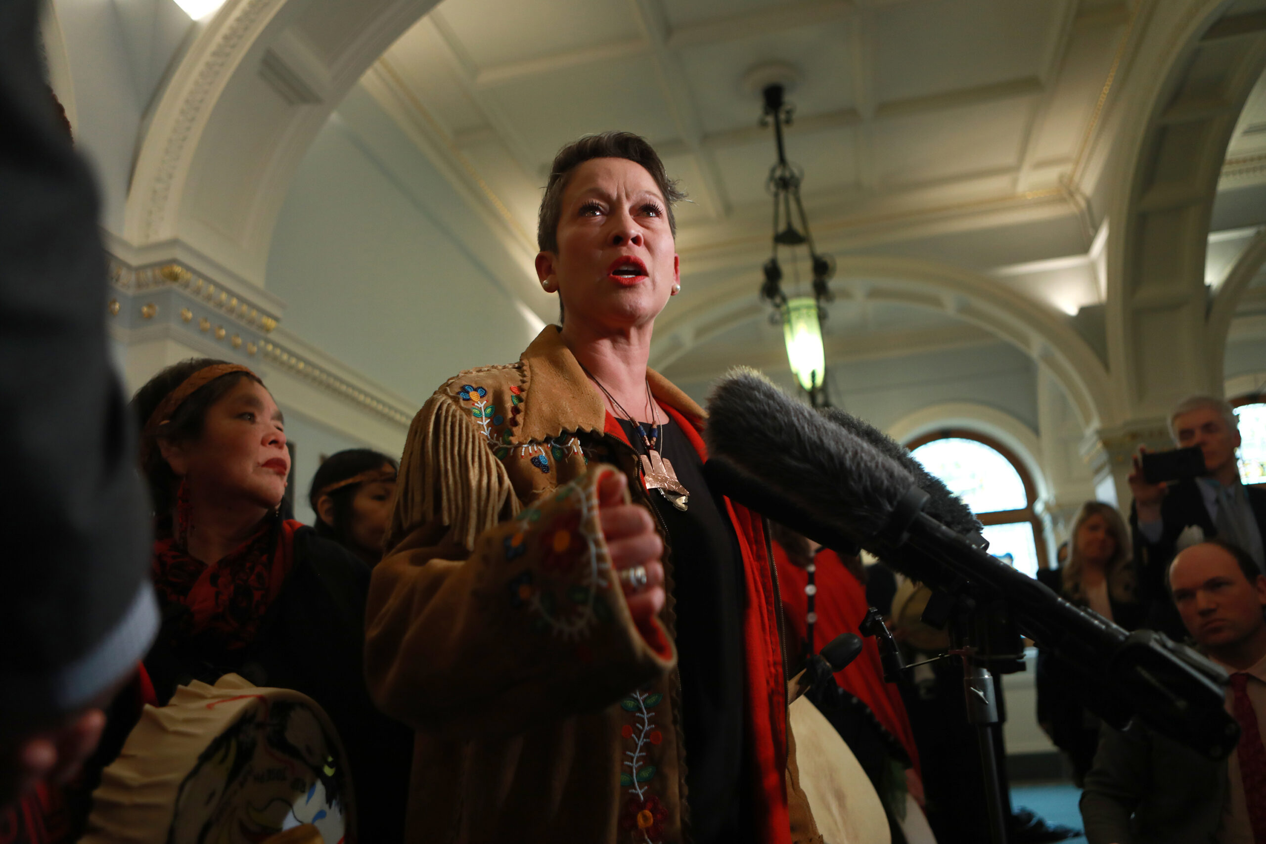 Melanie Mark parle devant des microphones dans une salle élégante avec des plafonds à caissons et des arcades. Elle est entourée de sympathisants autochtones. Elle tient un tambour plat et large à la hanche et est vêtue d’une veste en cuir fauve avec des glands et des fleurs vertes, roses et rouges peintes sur les poignets, et d’une chemise noire. Elle porte un ornement en bronze sur une chaîne en perles autour du cou.
