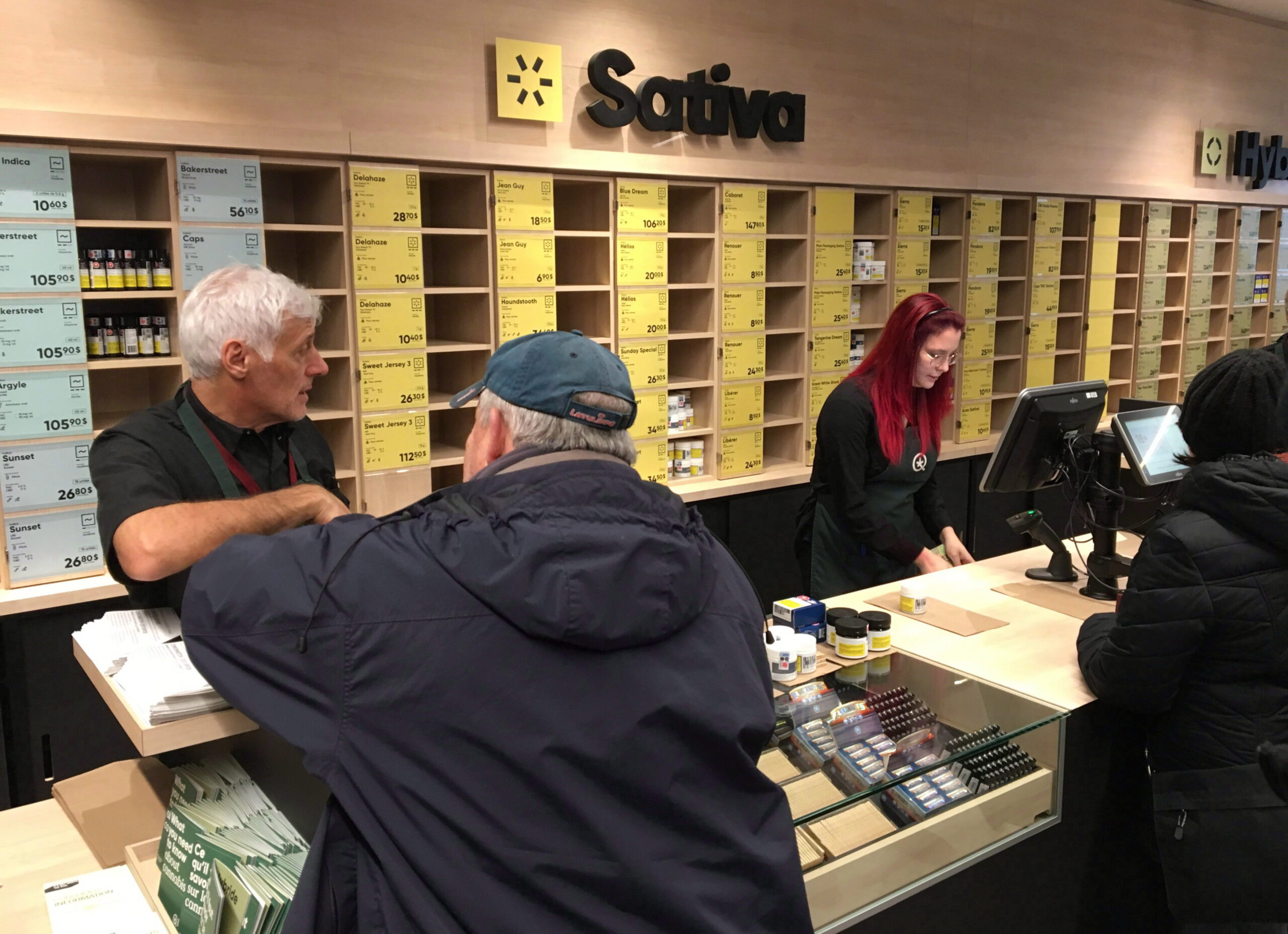Trois clients se tiennent devant un long comptoir où deux employés les servent derrière des ordinateurs. Une petite sélection de produits est présentée sur le comptoir. Le mur du fond est rempli d’alvéoles où sont rangés des petits produits. Plusieurs sont vides. Au-dessus des casiers, on peut lire le mot « sativa » en grosses lettres noires.