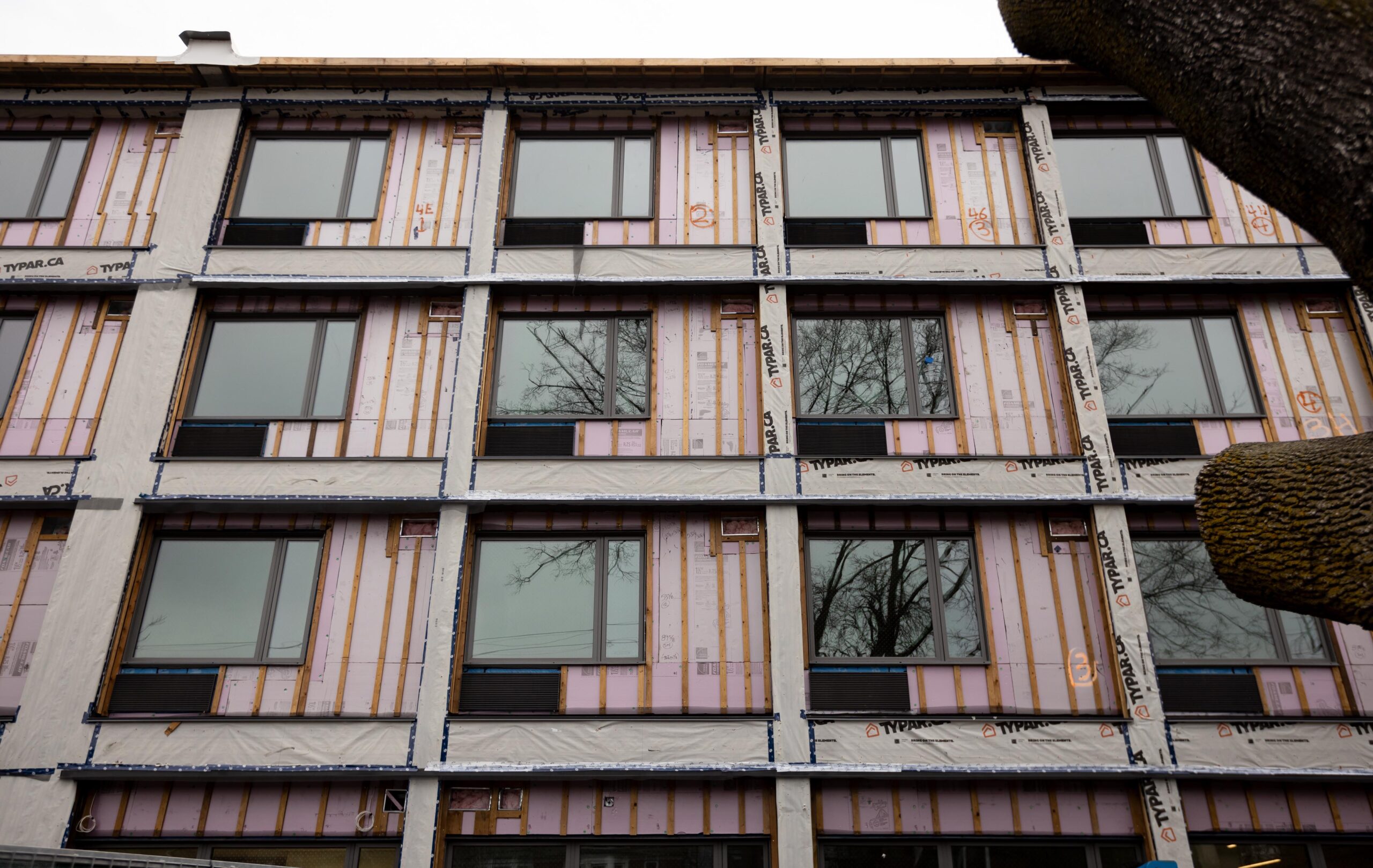 Close-up of the building, which has large square windows with a space underneath them for air-conditioning units. Pink sheets of insulation and an off-white membrane are visible on the exterior walls. 