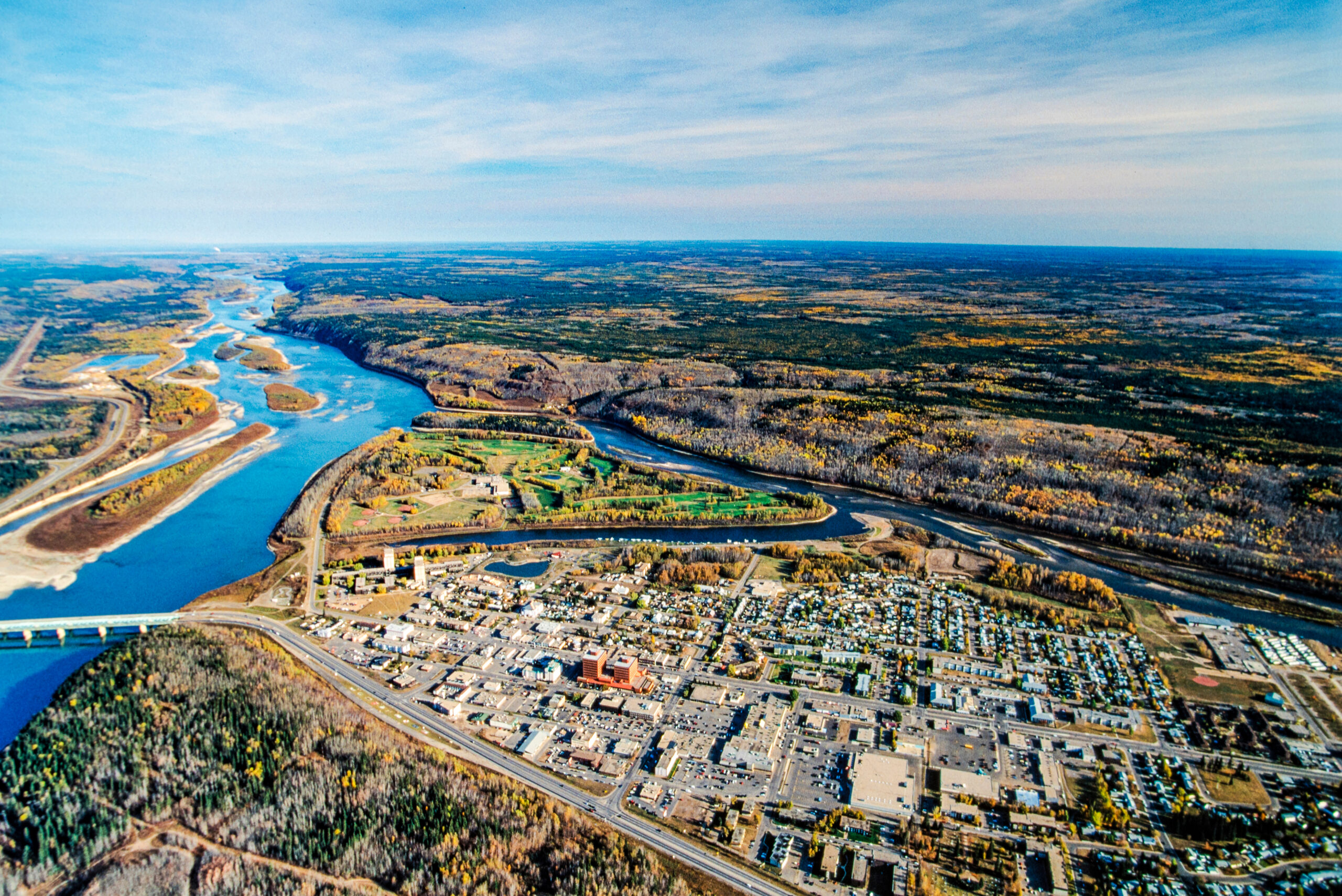 From the air, an industrial sector is bordered by a highway and two rivers, with a treed landscape that goes to the horizon. 