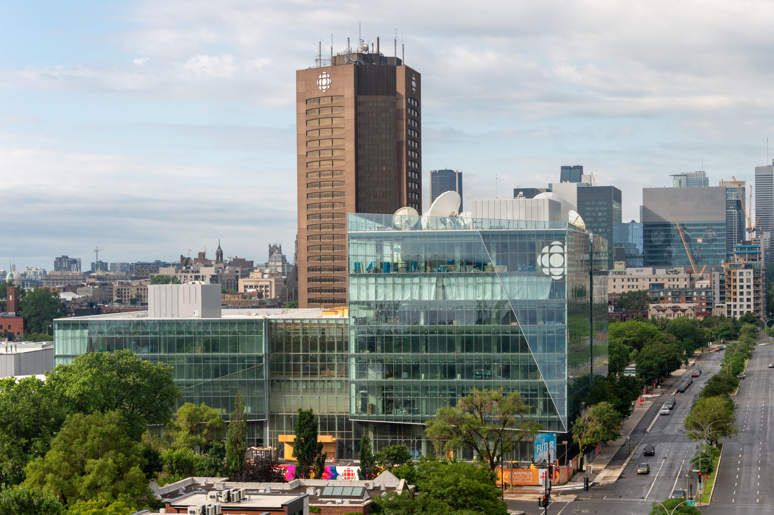 Shot from about five storeys up, the two buildings sit on a tree-lined city block lined with a major boulevard. The glass building has two parts, one about eight floors high, the other about six floors high. The tower in behind is about 25 storeys high and is made of brown brick.