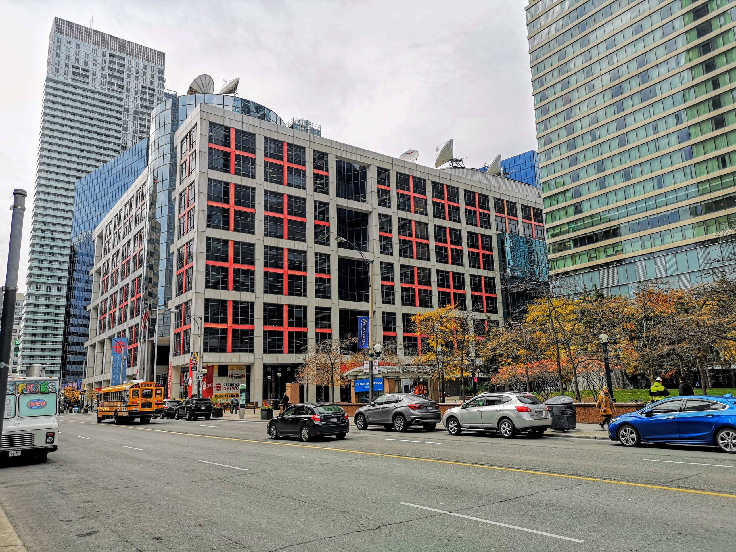 The building is about nine storeys high, and has a cross-hatch pattern of red panels that form a series of crosses on each floor. It’s situated on a busy street and surrounded by glass towers. Trees and parked cars line the street.