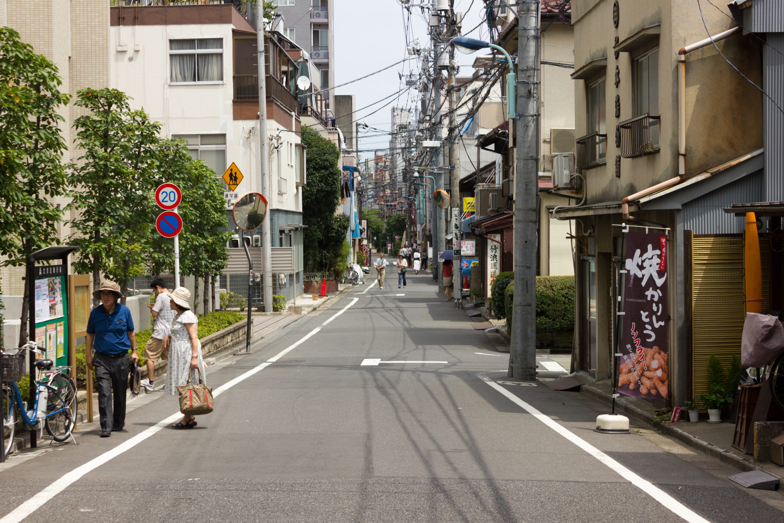 A one-lane street with no cars, parked or otherwise, is flanked by low-rise buildings on both sides. A few people are walking along the side.