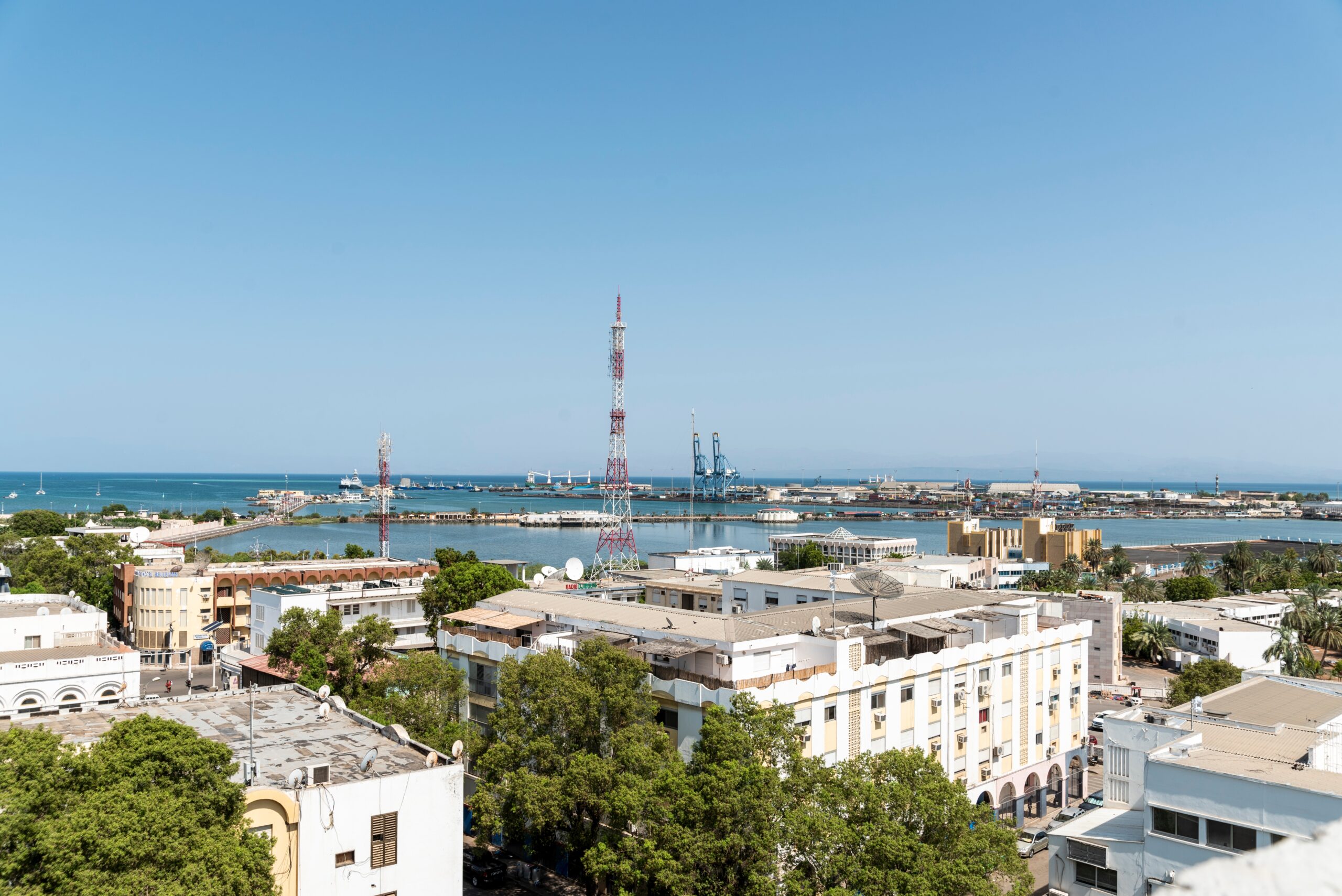 A working port with two cranes sits on the edge of a downtown area with lowrise, white-stone buildings. A lot of green trees line the streets. 