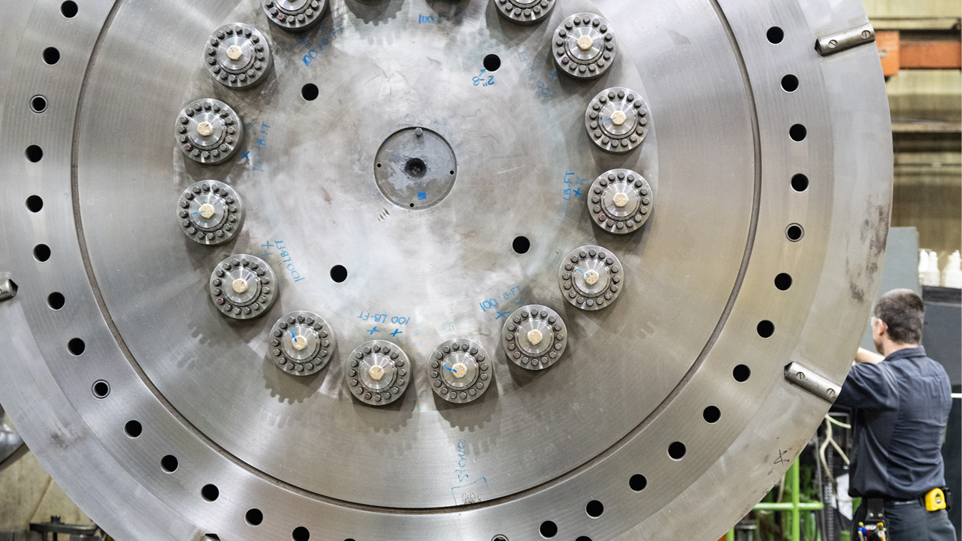 A man works on a shiny large steel wheel more than twice his size.