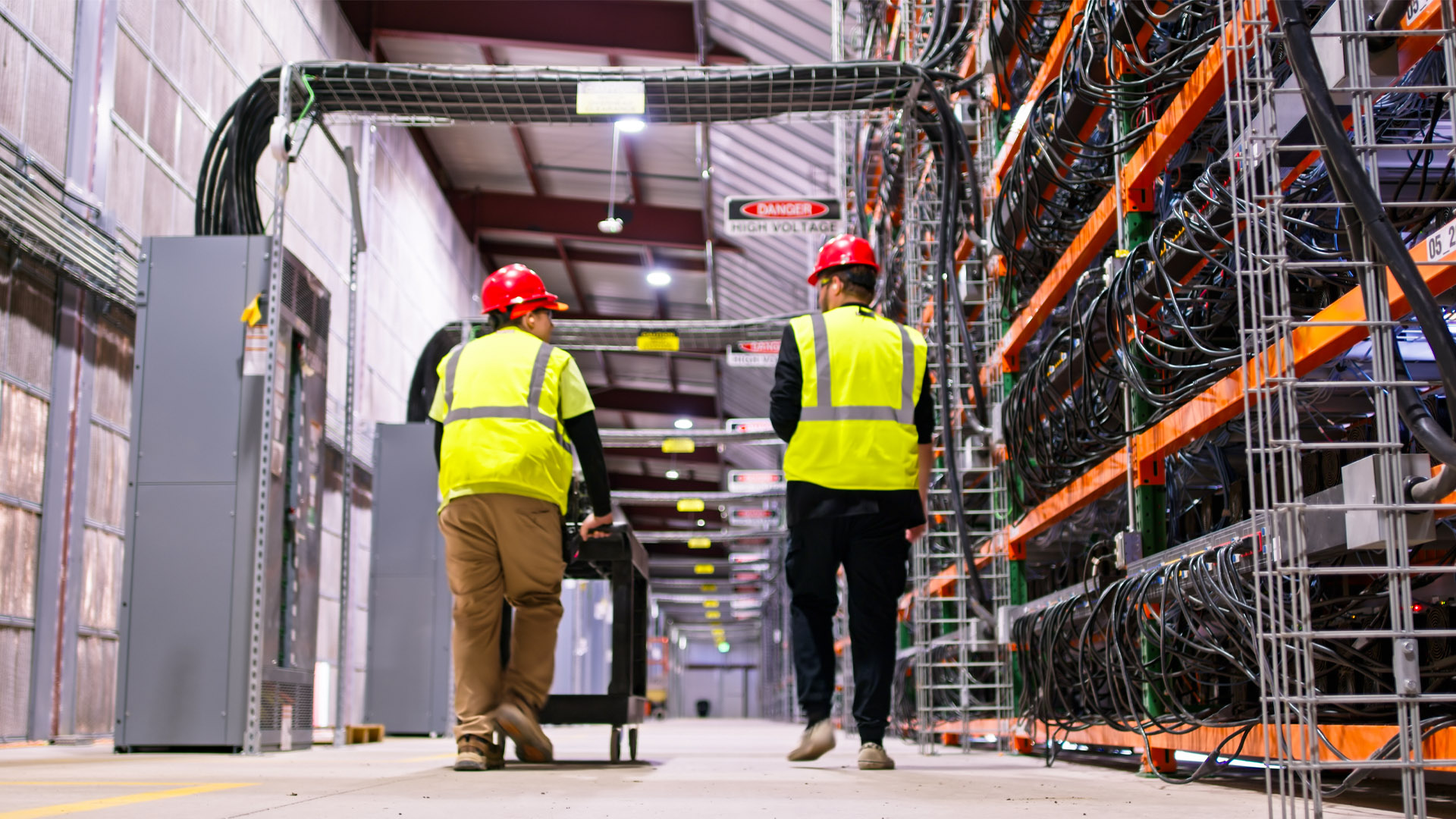 Two technicians wearing yellow high visibility coats and hard hats walking past racks of equipment in a data centre.