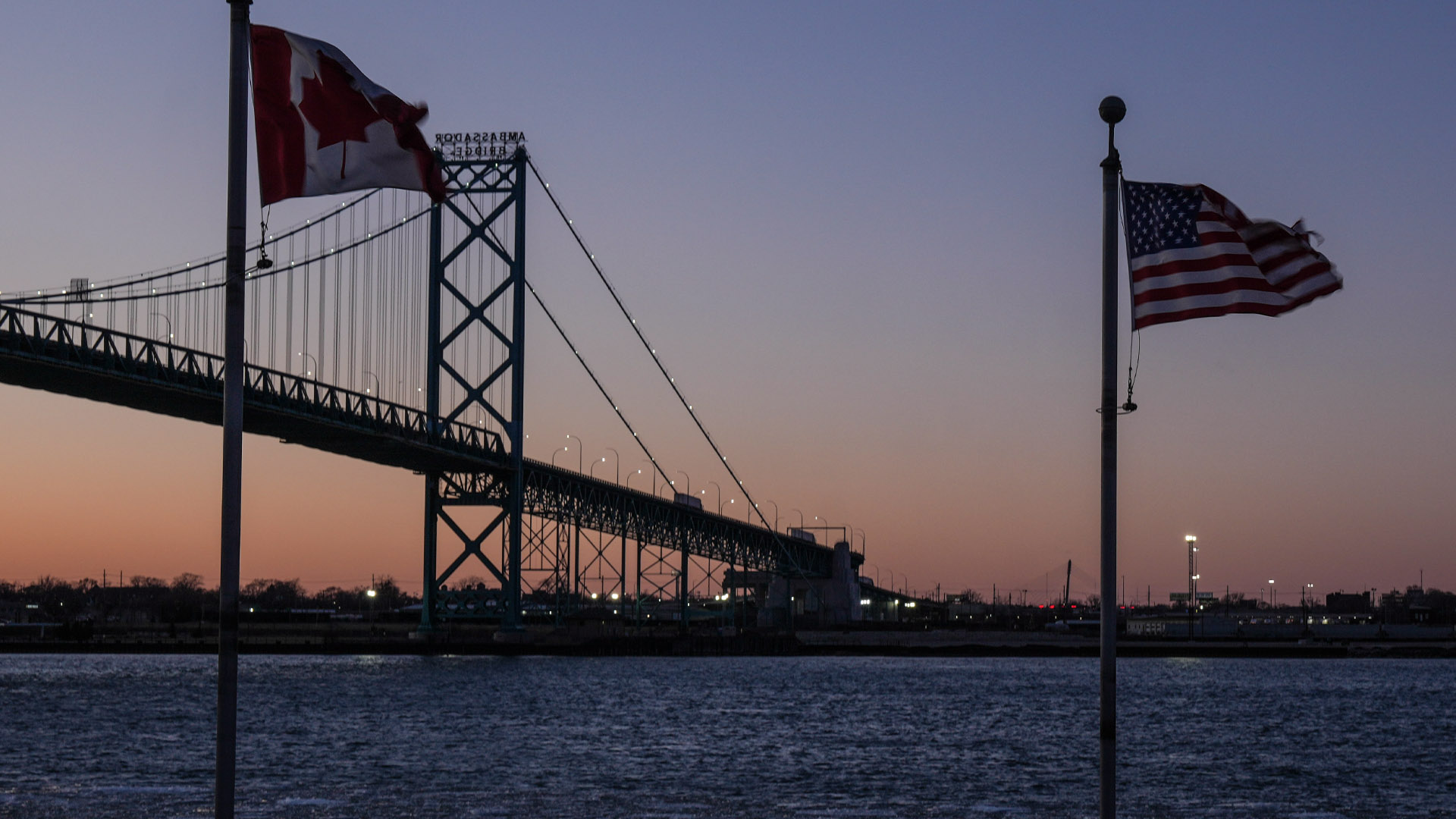 The Canadian and U.S. flags are in the foreground with the Ambassador bridge in the background before a pink sunset.