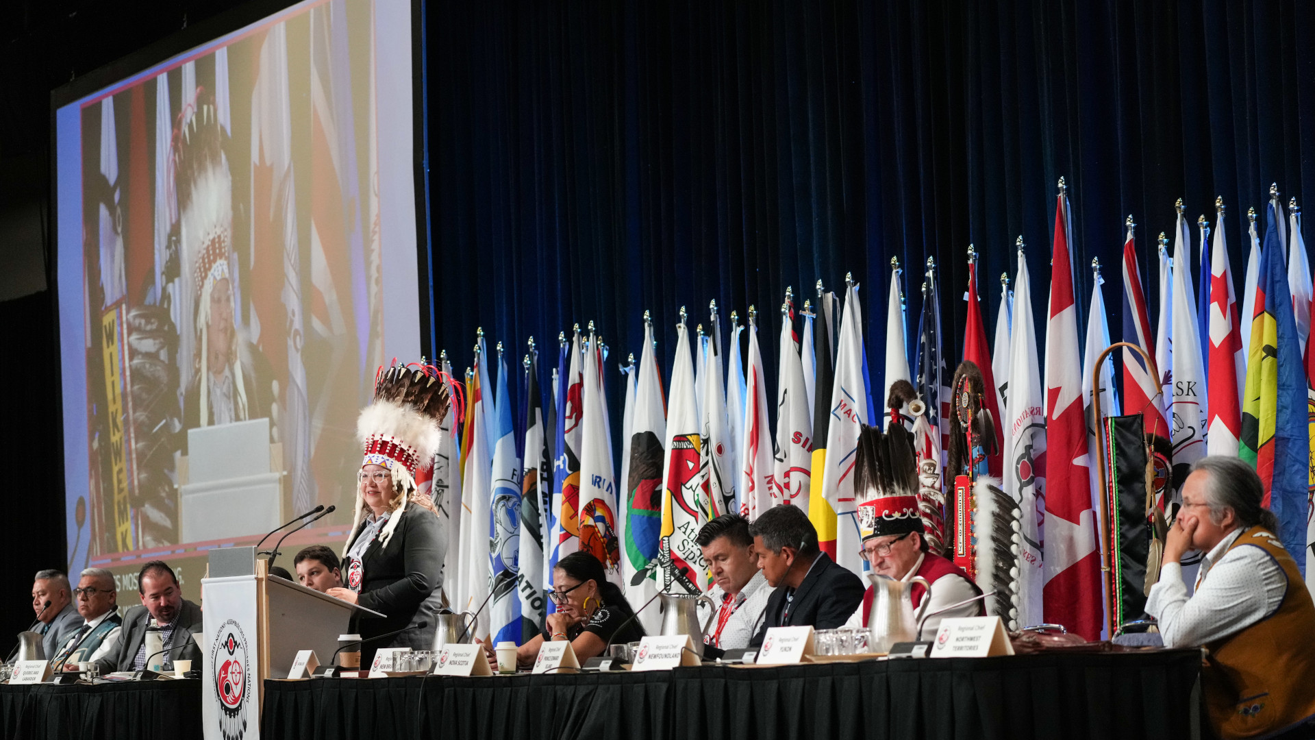Indigenous National Chief Cindy Woodhouse speaking at podium during an assembly, surrounded by First Nations representatives and flags.