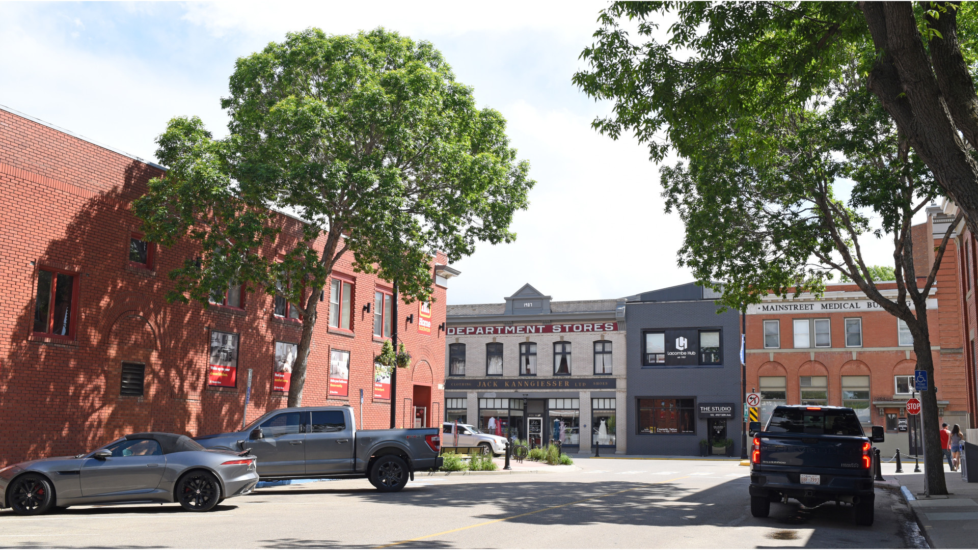 Two downtown streets intersect, with two mature trees and four parked cars. No cars, no people. A medical clinic and department store have storefronts.