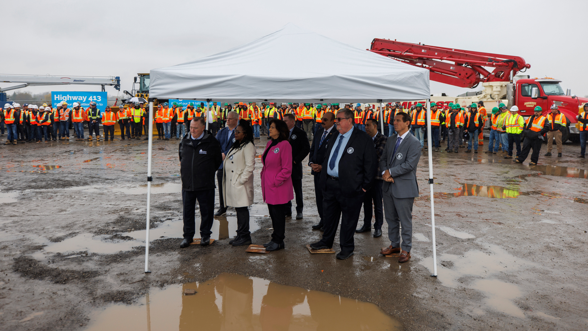 Nine people stand under a white canvas tent surrounded by mud and puddles on a cloudy day. A long line of men in fluorescent orange and lime-green work vests and hardhats is behind them along with a couple of crane trucks.
