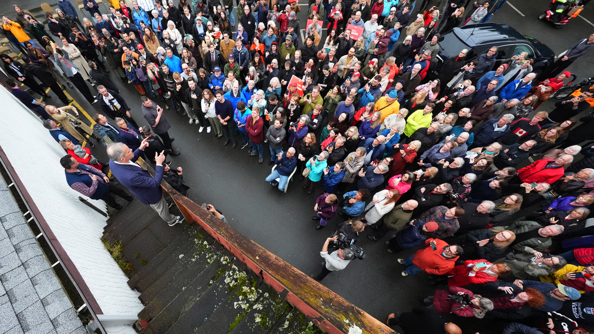 An overhead view of Mark Carney standing on steps speaking to dozens of supporters.