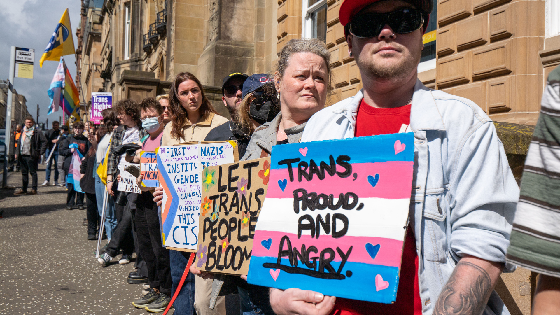 Manifestation pour les droits des personnes transgenres devant un bâtiment. Des manifestants tiennent des pancartes aux messages pro-trans comme « Trans, proud, and angry ». Drapeaux trans et LGBTQ+, ambiance pacifique, chien au premier plan.