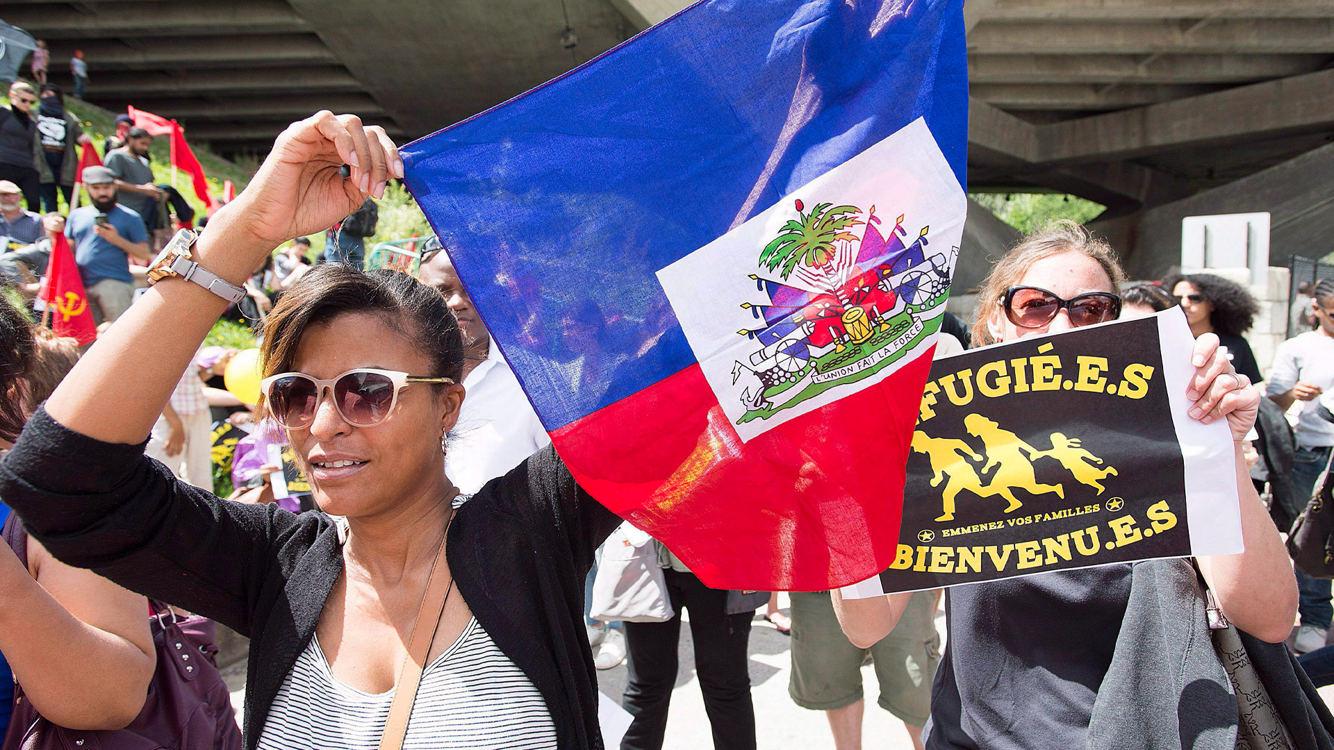 Des manifestantes tiennent un drapeau haïtien et une pancarte « Réfugié.e.s bienvenu.e.s » lors d’un rassemblement.