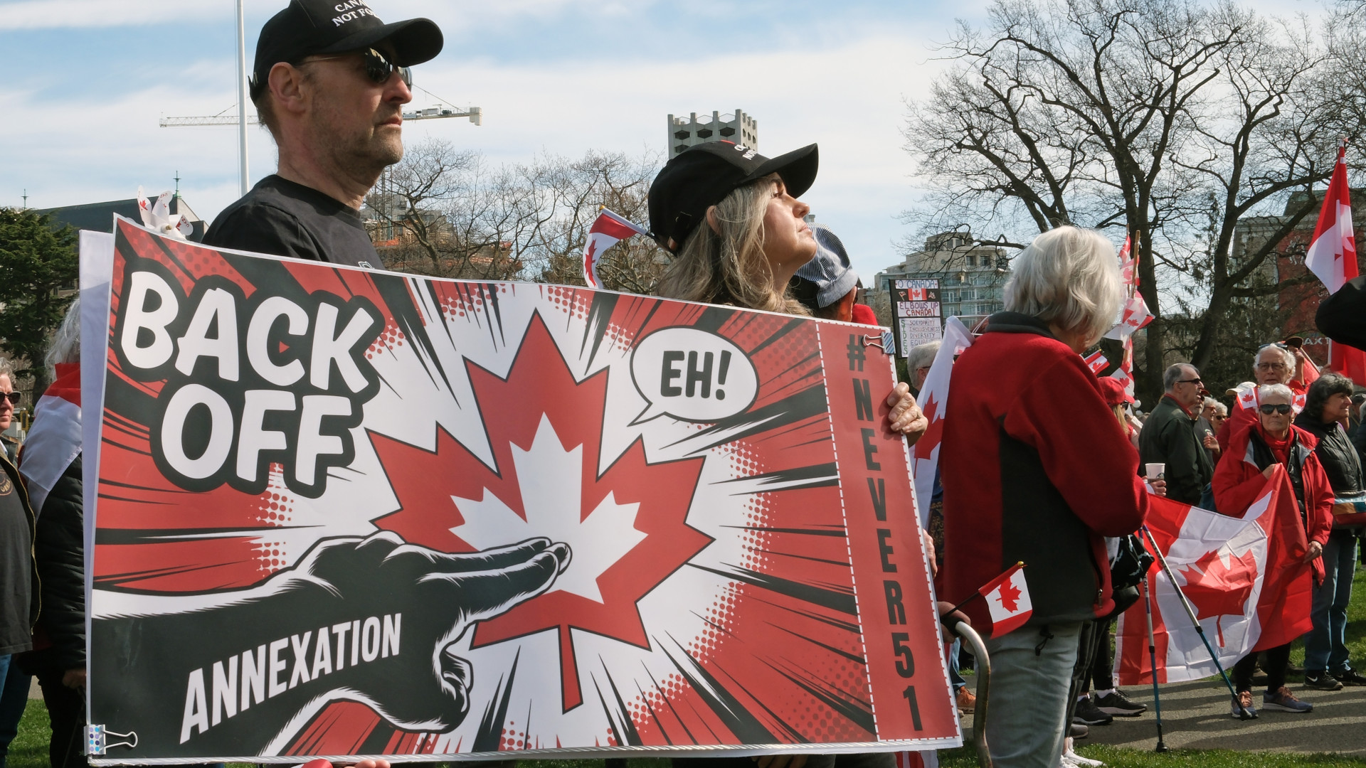 A couple holds a sign with a Maple Leaf that says “back off eh” amid a crowd of protesters with flags.