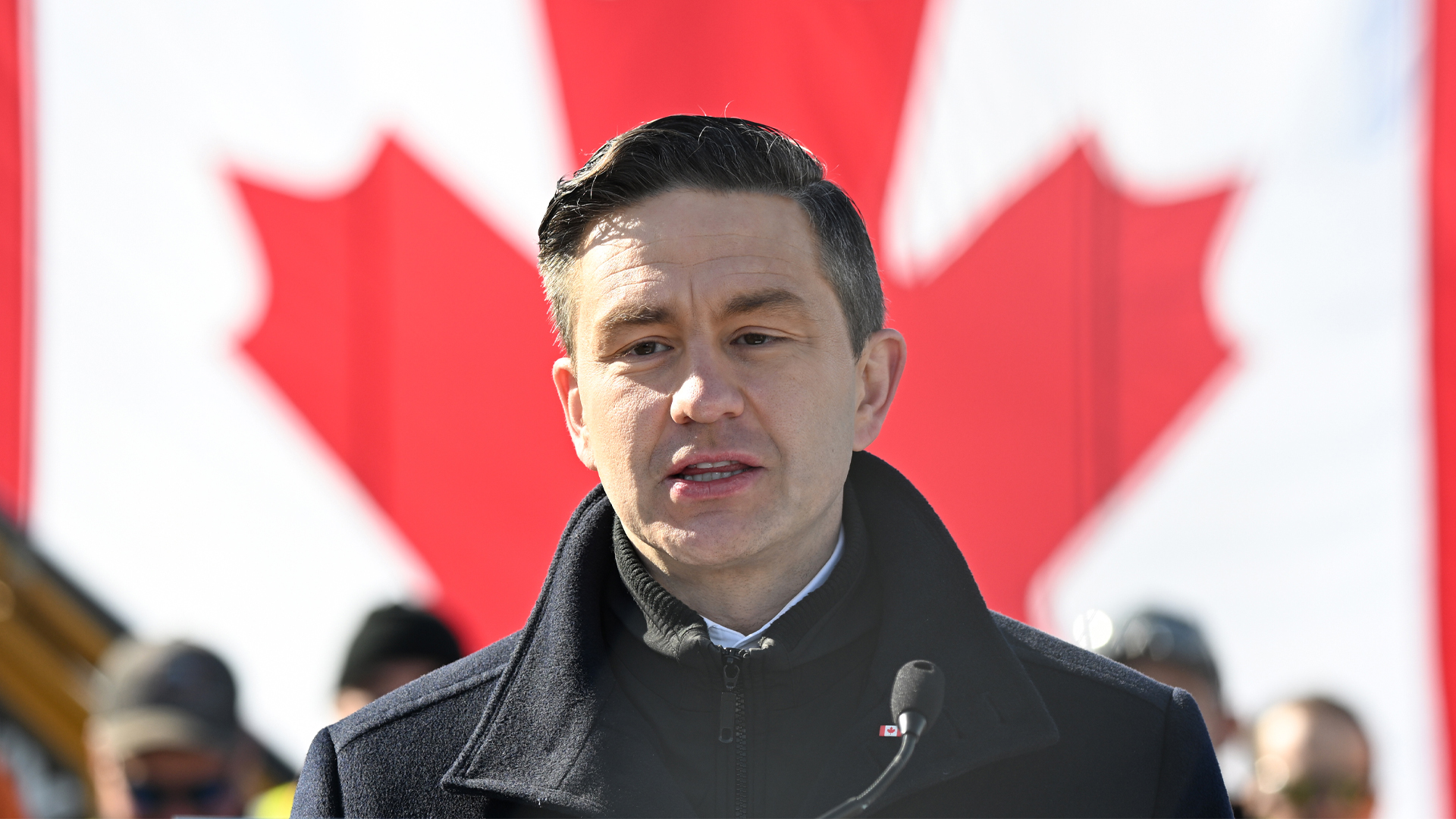 Pierre Poilievre at a microphone standing in front of a Canadian flag.