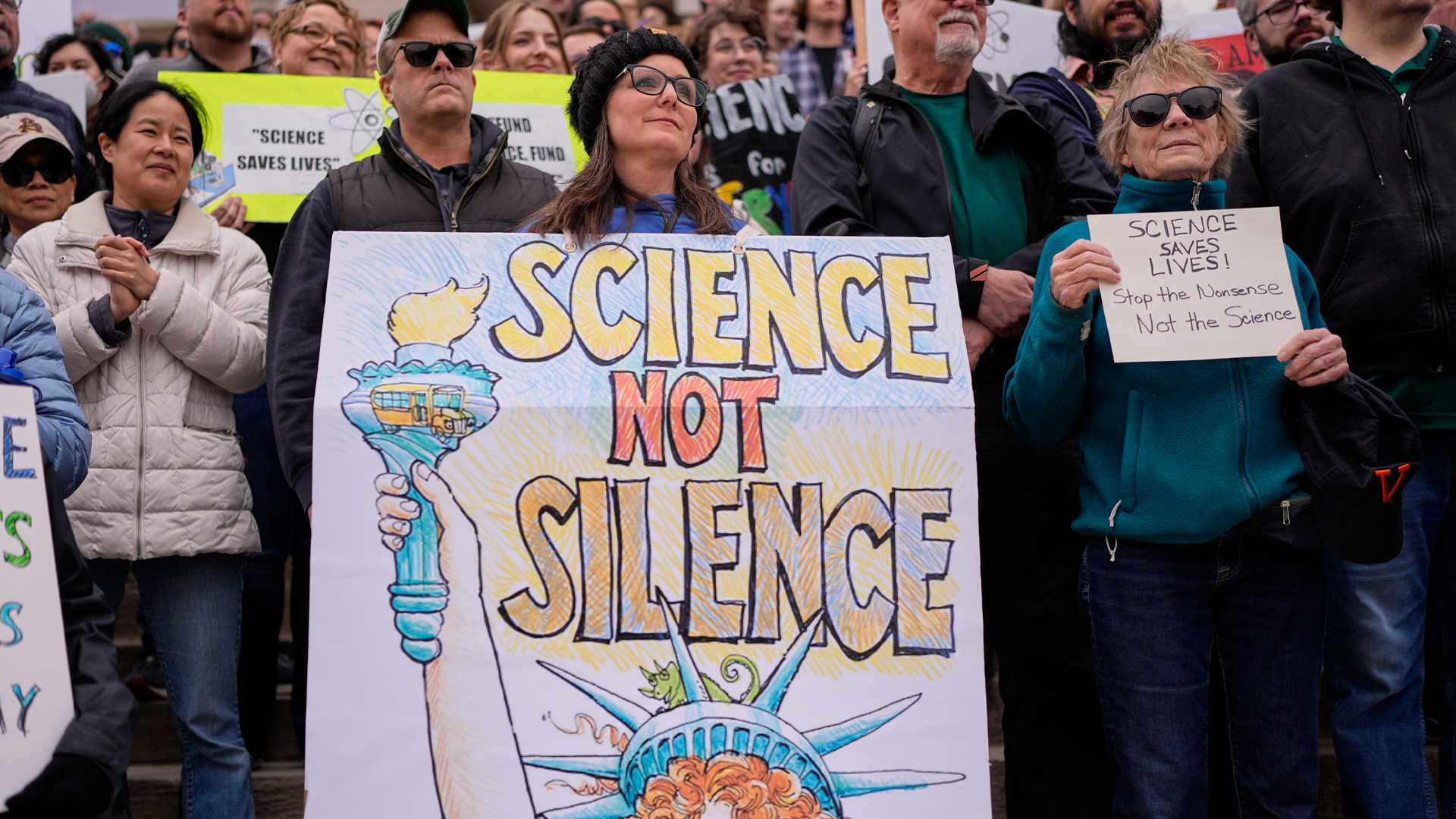 Protesters stand behind a banner with a hand-drawn Statue of Liberty and with the slogan “Science not silence” written on it.